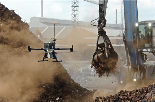 Eine Drohne fliegt über einem Recycling- oder Schrottplatz und misst offenbar Umwelt- oder Luftdaten, während daneben ein Bagger mit Greifarm Metall- oder Schrottmaterial bewegt. Staub wirbelt auf, im Hintergrund sind Industriegebäude und Anlagen zu sehen.